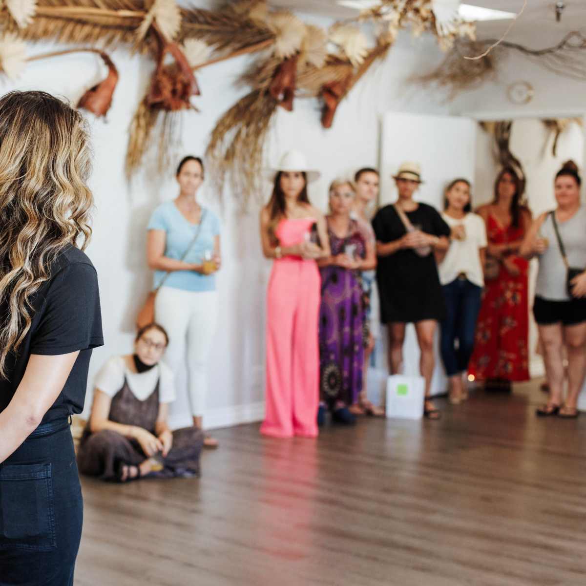 A woman with long hair stands facing a group of people in a decorated room with plants and artistic elements on the walls.
