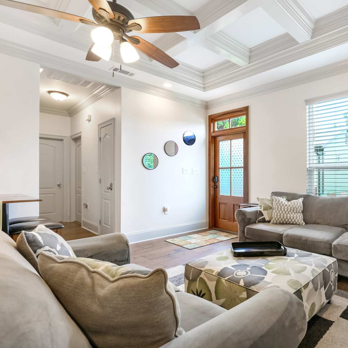 A bright living room with a ceiling fan, gray sofas, a decorative ottoman, and a doorway leading outside.