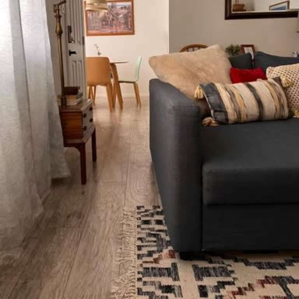 A living room features a gray sofa adorned with various pillows, alongside a side table and a lamp, leading towards a dining area with a wooden table and chairs in the background.