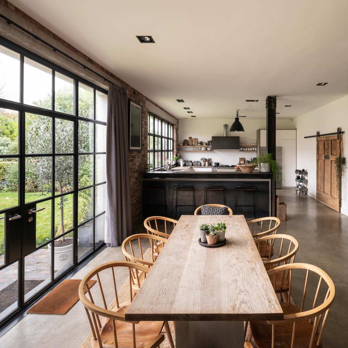 A modern kitchen and dining area with a large wooden table, surrounding chairs, and large windows that open to a garden.