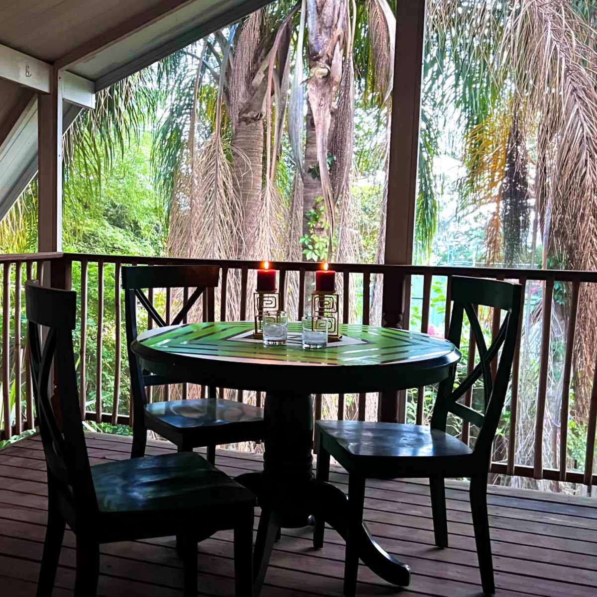 A small wooden dining table with four chairs is set on a porch surrounded by tropical greenery.