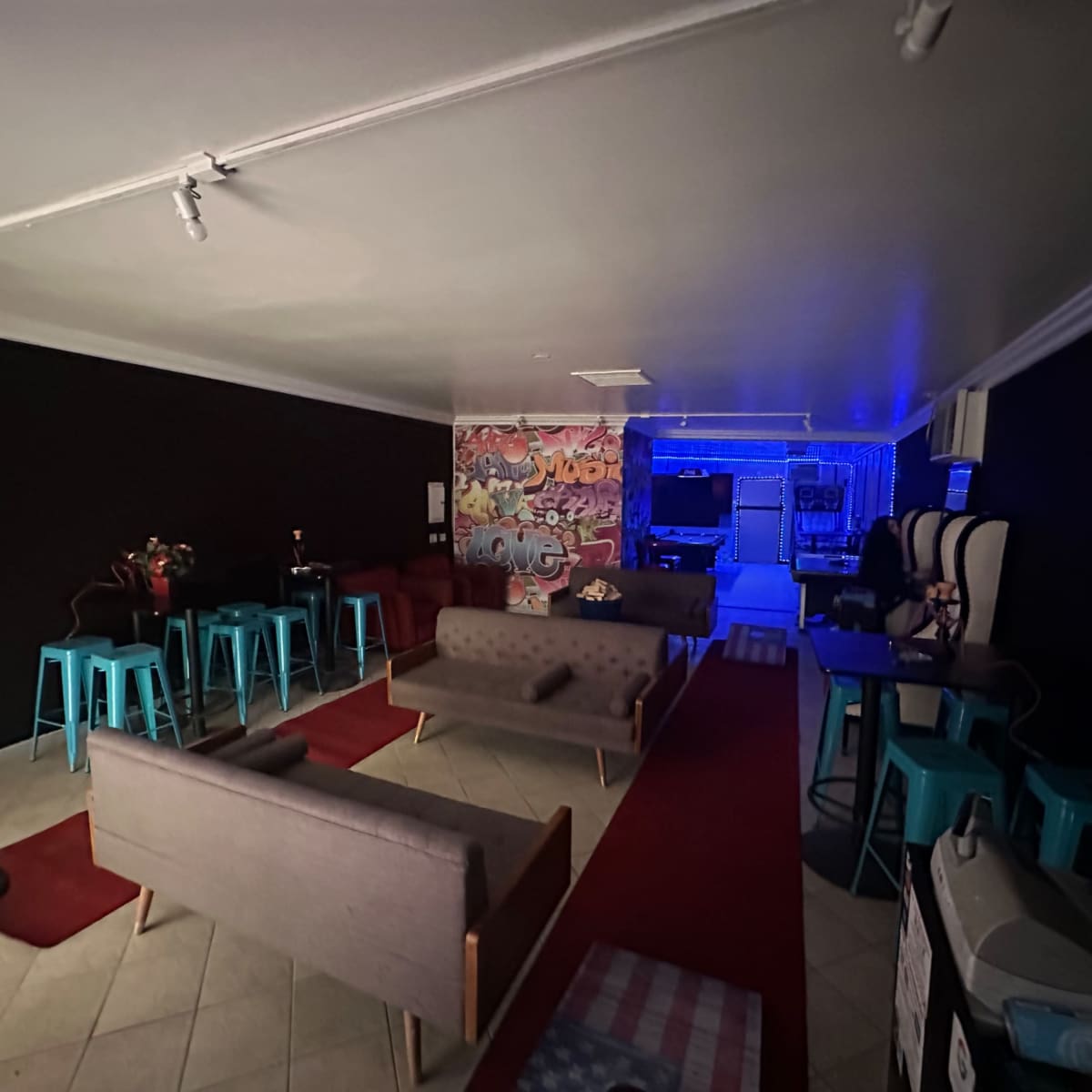 A dimly lit lounge area with gray couches, teal stools, and a wall featuring colorful graffiti.