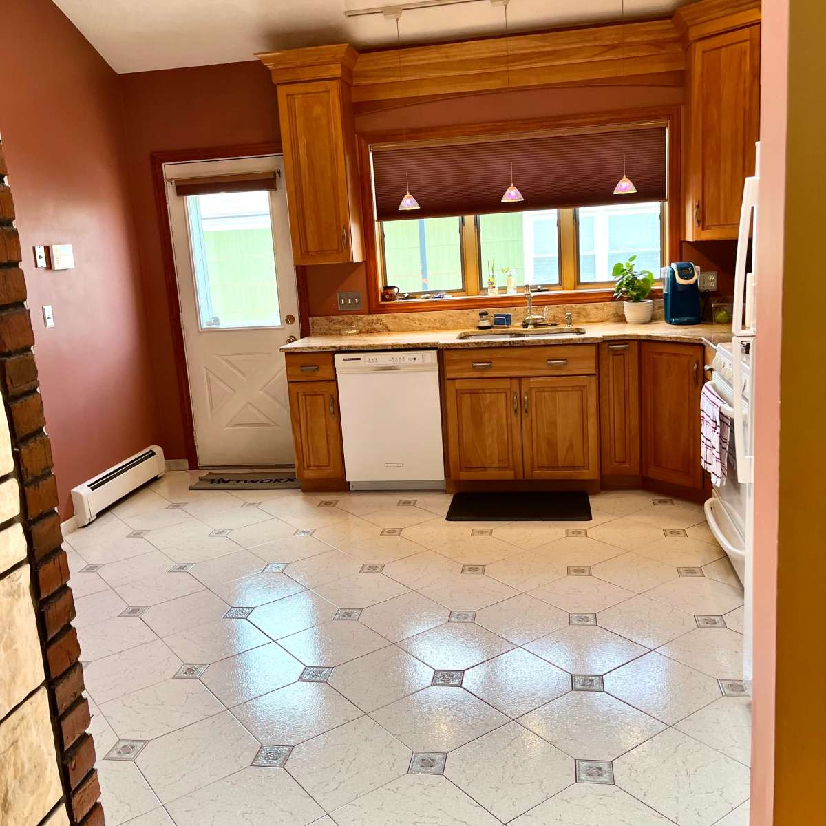 A kitchen features wooden cabinetry, a dishwasher, and a door leading outside, with tiled flooring and overhead lighting.