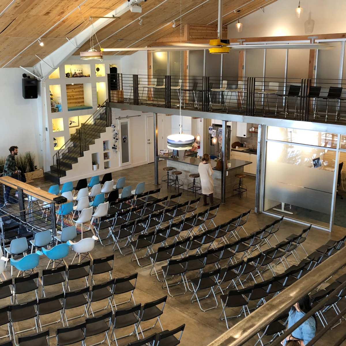 An interior space designed for gatherings, featuring rows of black folding chairs, a stage with a wooden bar, and a multi-level layout with large windows and a modern aesthetic.