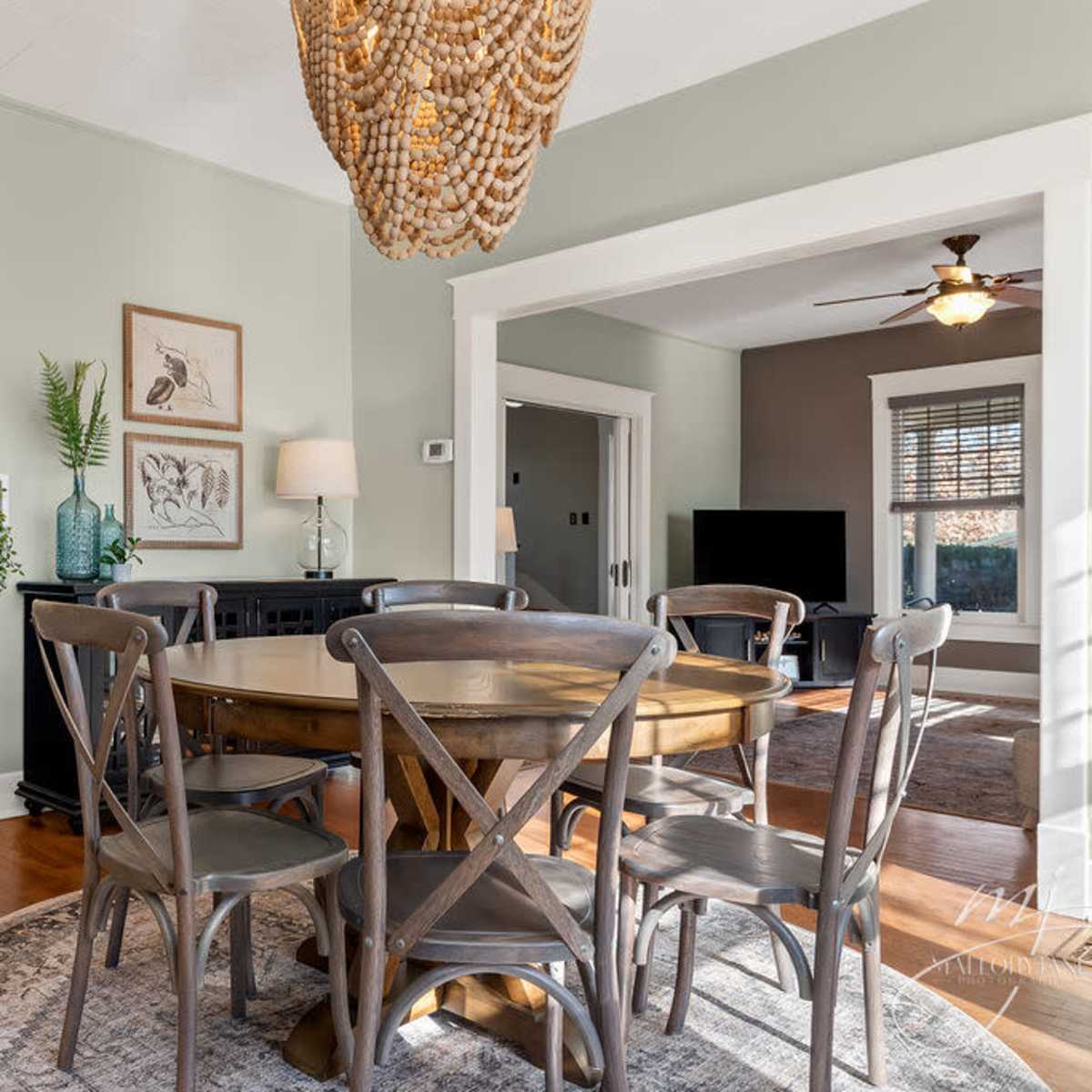 A dining room features a wooden table surrounded by gray chairs, with a large light fixture hanging overhead and doorways leading to adjacent rooms.