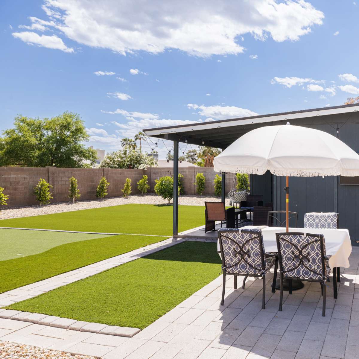 A patio area features a white umbrella over a dining table surrounded by six chairs, with artificial turf and landscaped greenery in the background.