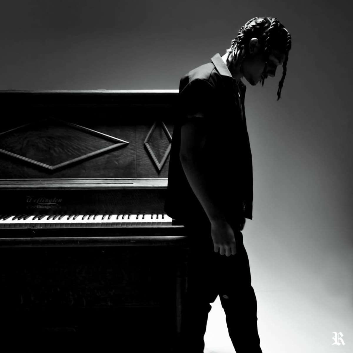 A person stands beside an upright piano in a dimly lit space, gazing downward.
