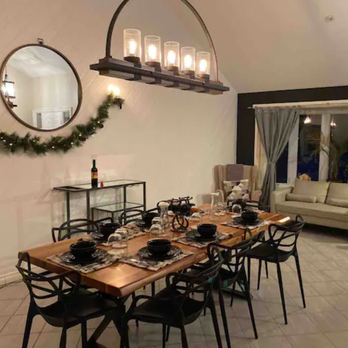 A dining area features a large wooden table set for a meal, surrounded by black chairs, with a cozy living space visible in the background.