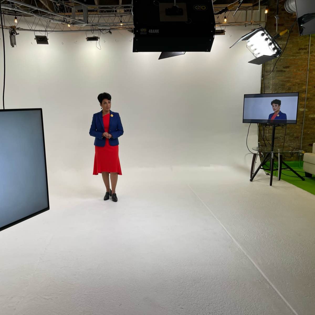 A person in a red dress and blue jacket stands in a brightly lit studio, flanked by two monitors displaying her image.
