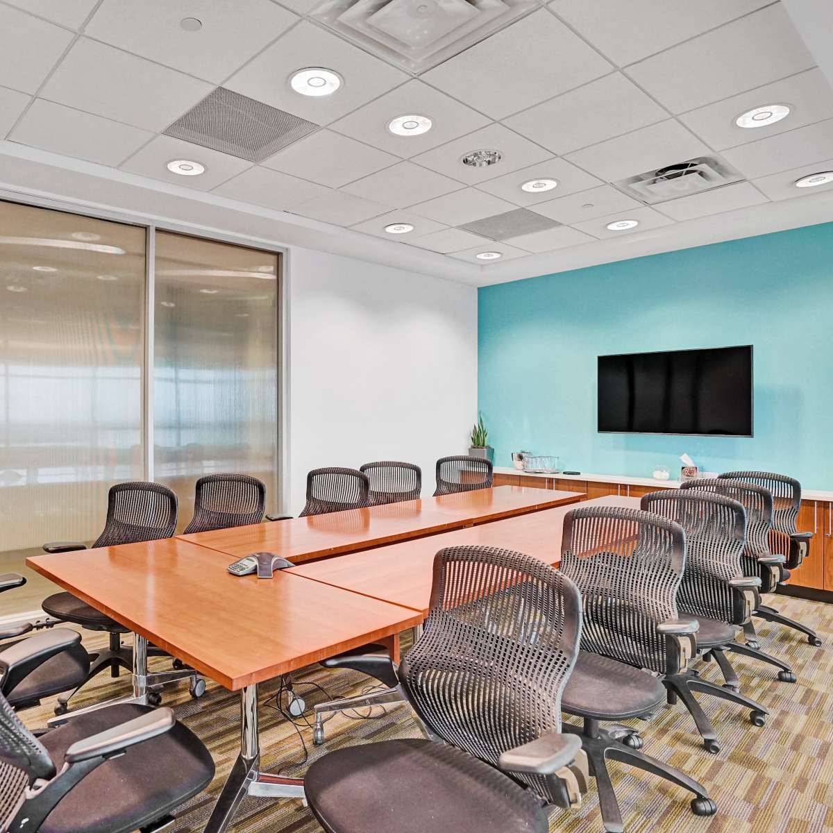 A modern conference room features a large wooden table surrounded by black mesh chairs, with a television mounted on the teal wall and large glass windows allowing natural light.