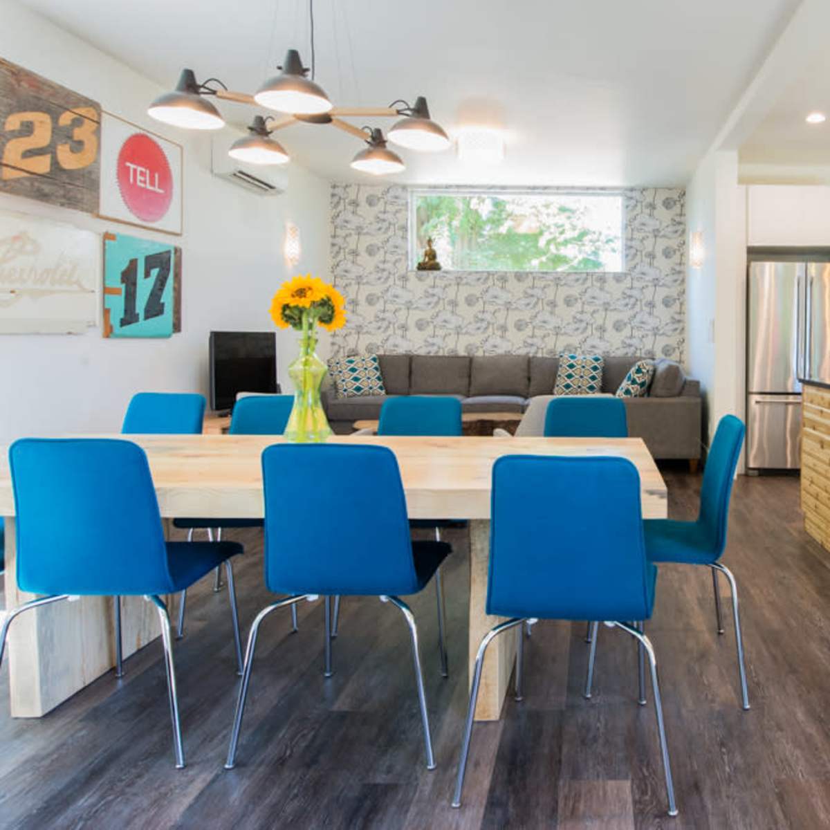 A modern open-concept living space features a dining area with a wooden table surrounded by blue chairs and a kitchen bar with stools.