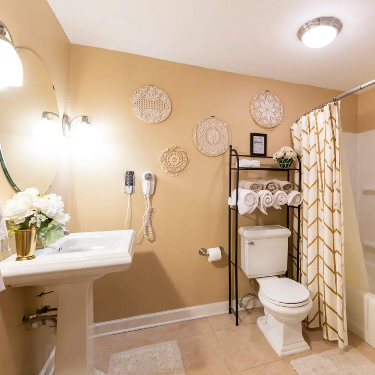 The bathroom features a sink with a mirror, a toilet, a bathtub with a shower curtain, and a shelving unit displaying neatly rolled towels.
