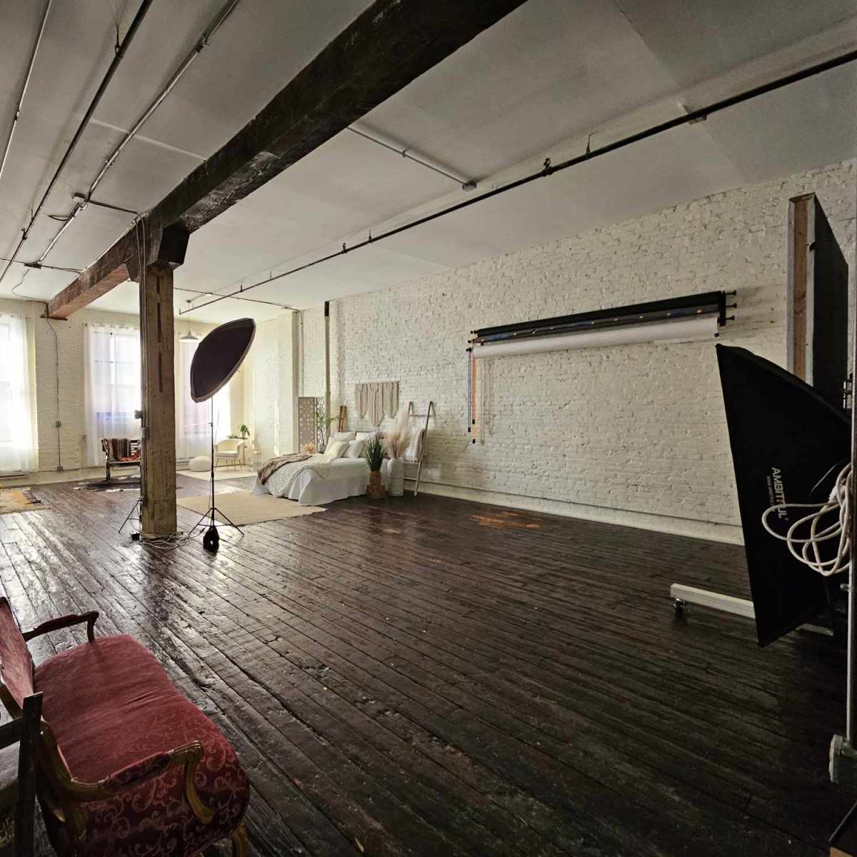 A spacious studio with wooden floors, white brick walls, large windows, and various furnishings, including a bed and photography equipment.