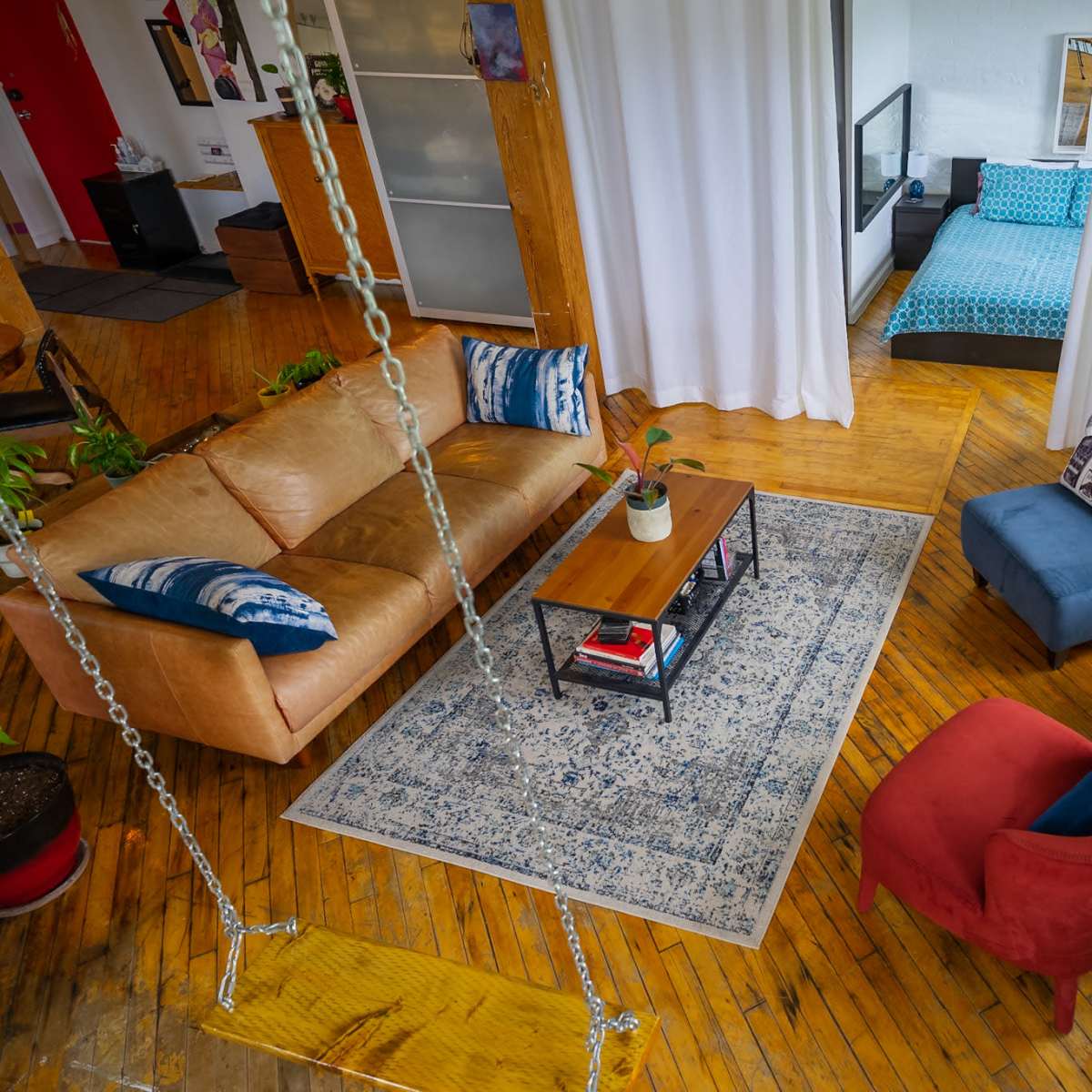 A spacious living area with a brown sofa, a blue armchair, a red chair, a coffee table on a patterned rug, and plants, with a bedroom visible in the background.