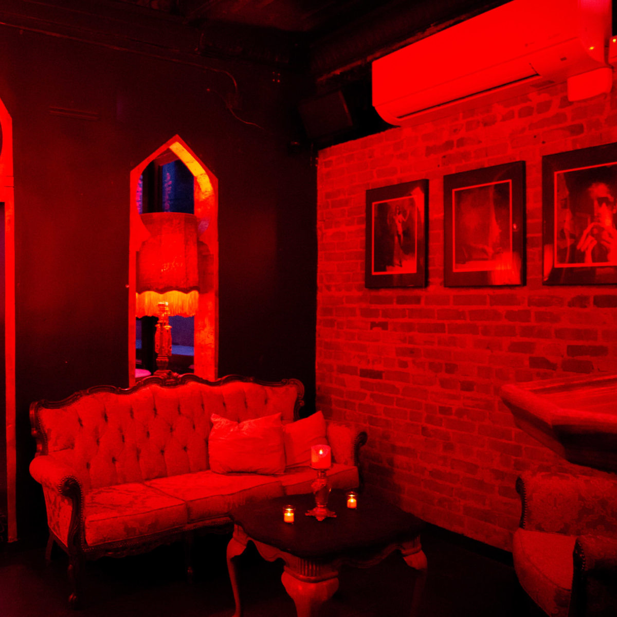 A dimly lit lounge with red lighting, featuring ornate archways, a vintage couch, and framed photographs on the walls.