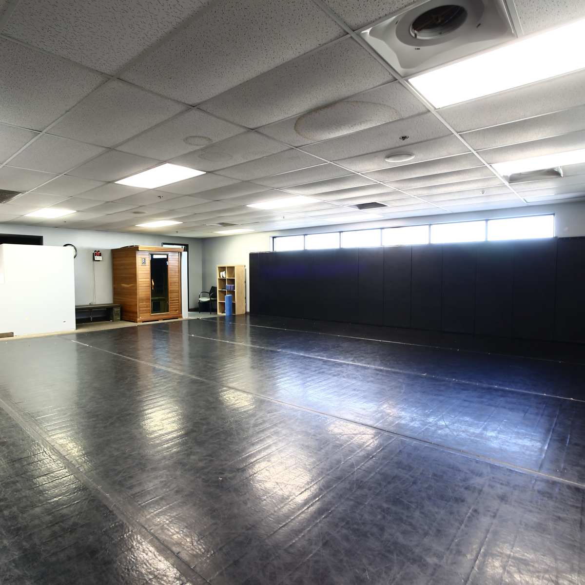 A large, empty room with black mats on the floor and padded walls, featuring bright overhead lights and large windows.