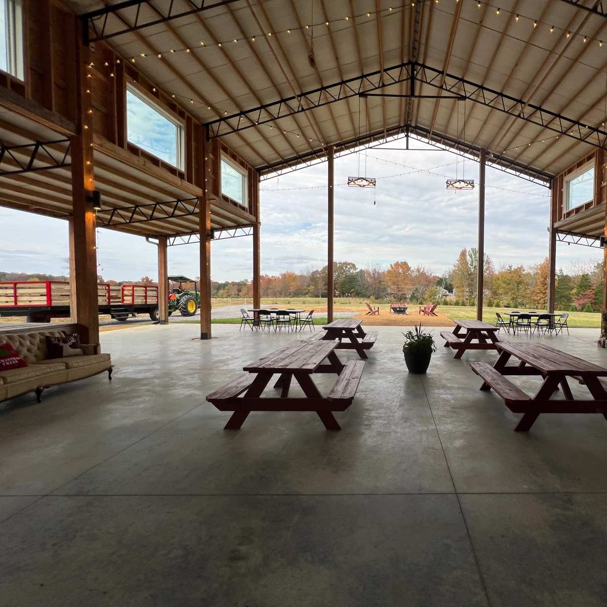 A spacious barn interior with wooden picnic tables, large windows, and a view of a landscaped outdoor area with a tractor and firewood stacks.