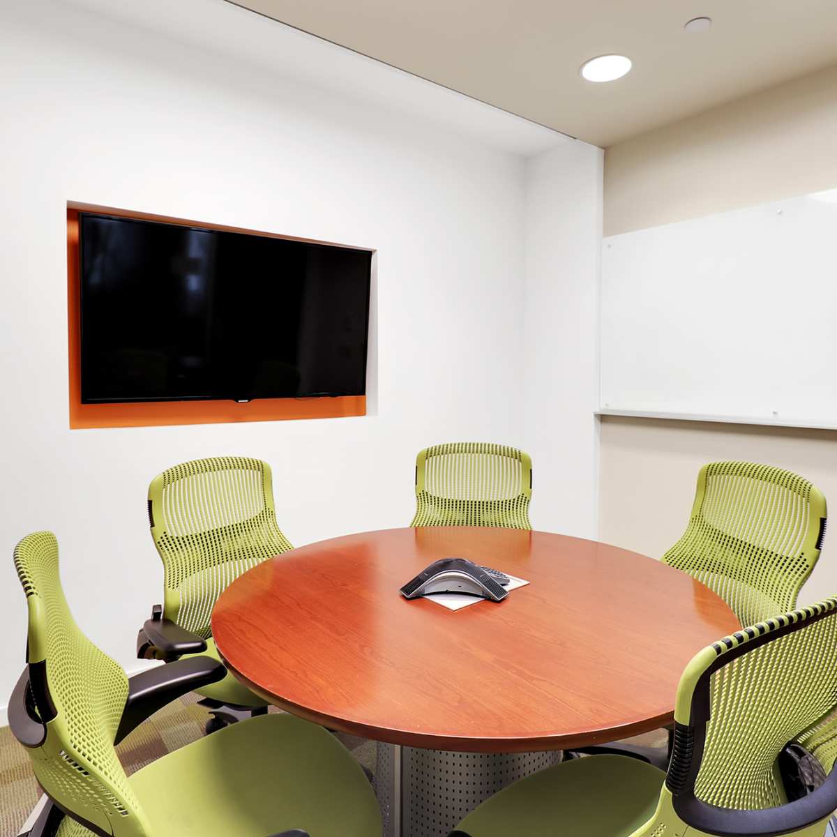 A conference room features a round wooden table with a speakerphone at the center, surrounded by four green chairs, along with a television mounted on the wall and a whiteboard nearby.