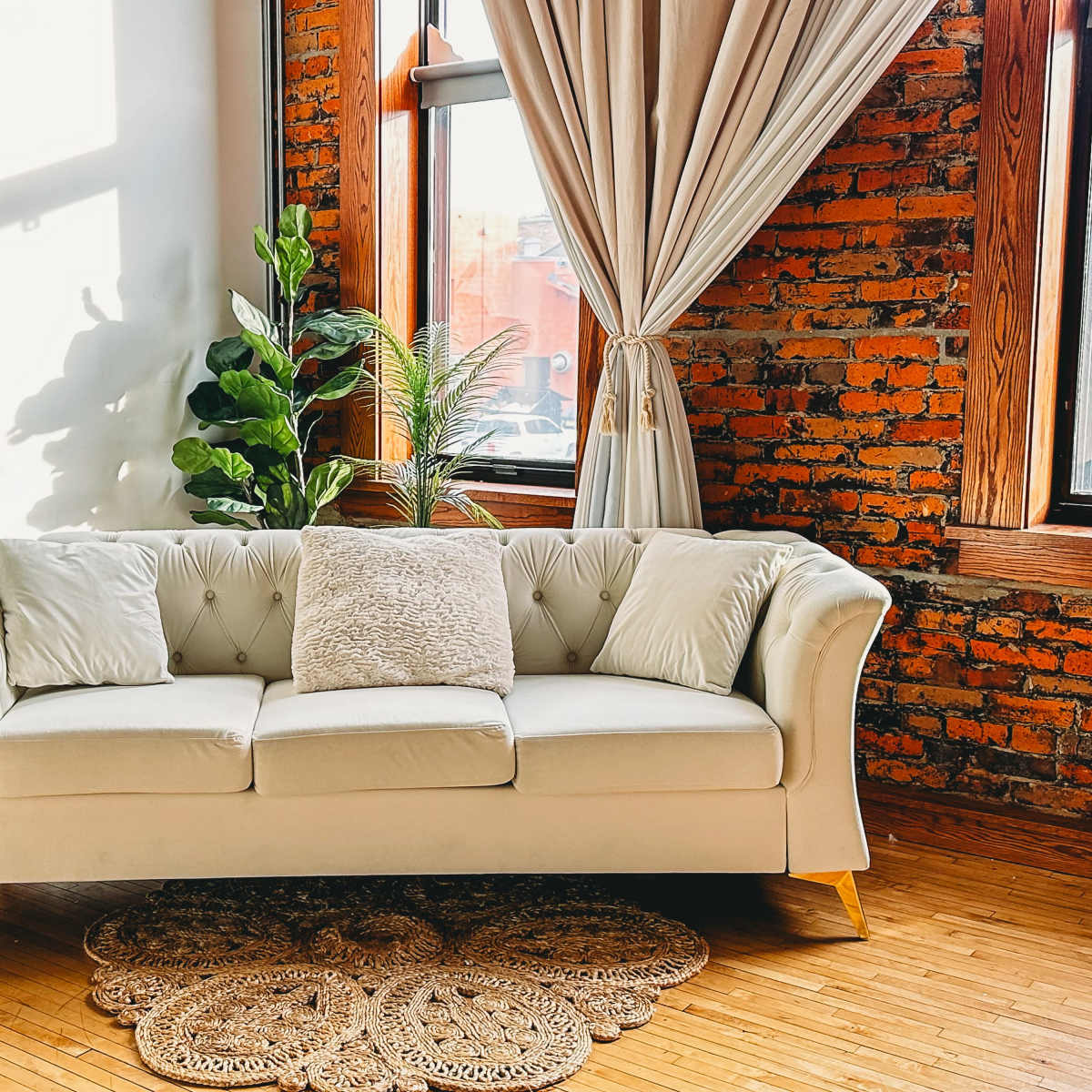 A light-colored chesterfield sofa with three pillows is positioned against a brick wall, complemented by a decorative plant and a woven rug on the wooden floor.