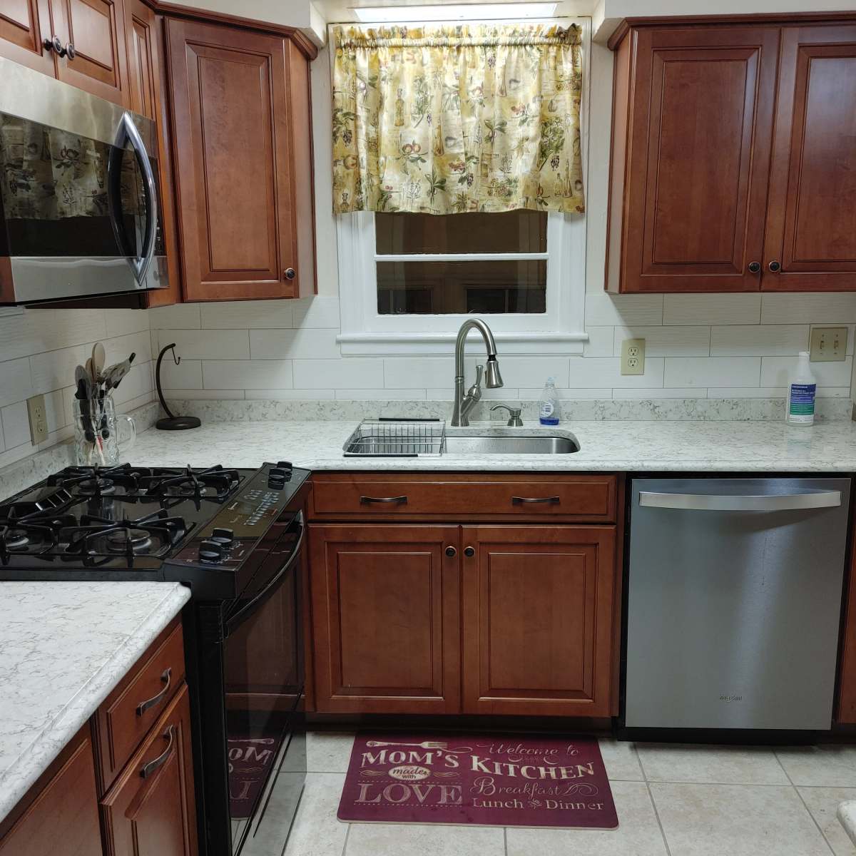 A kitchen with wooden cabinets, a sink beneath a window with a floral curtain, a stove, and a stainless steel dishwasher.