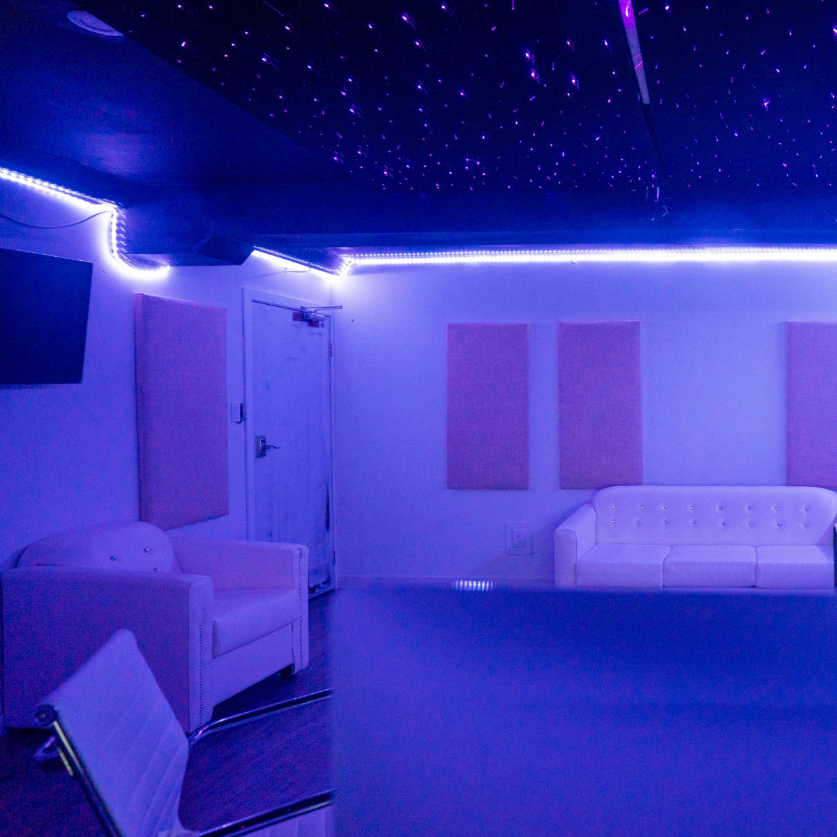 A modern, dimly lit studio space with a white couch, soundproof panels on the walls, and ambient purple lighting.