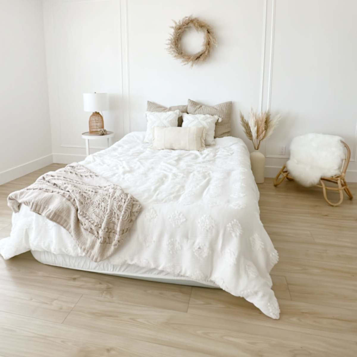 A bright, minimalist bedroom with a large bed covered in white bedding, a decorative wreath on the wall, and a cozy armchair in the corner.