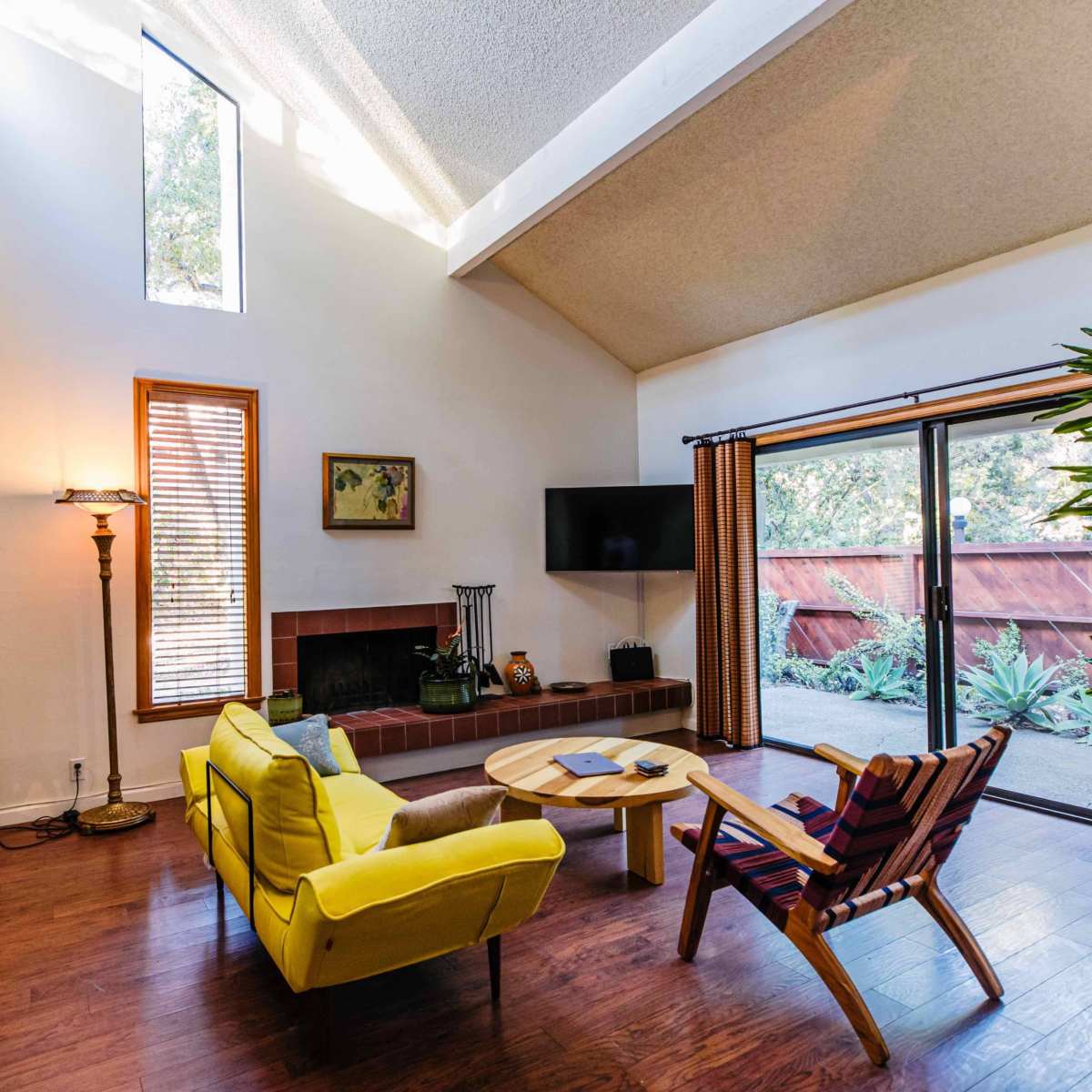 A bright living room features yellow sofas, a wooden coffee table, a bookshelf, and large sliding glass doors leading to an outdoor space.