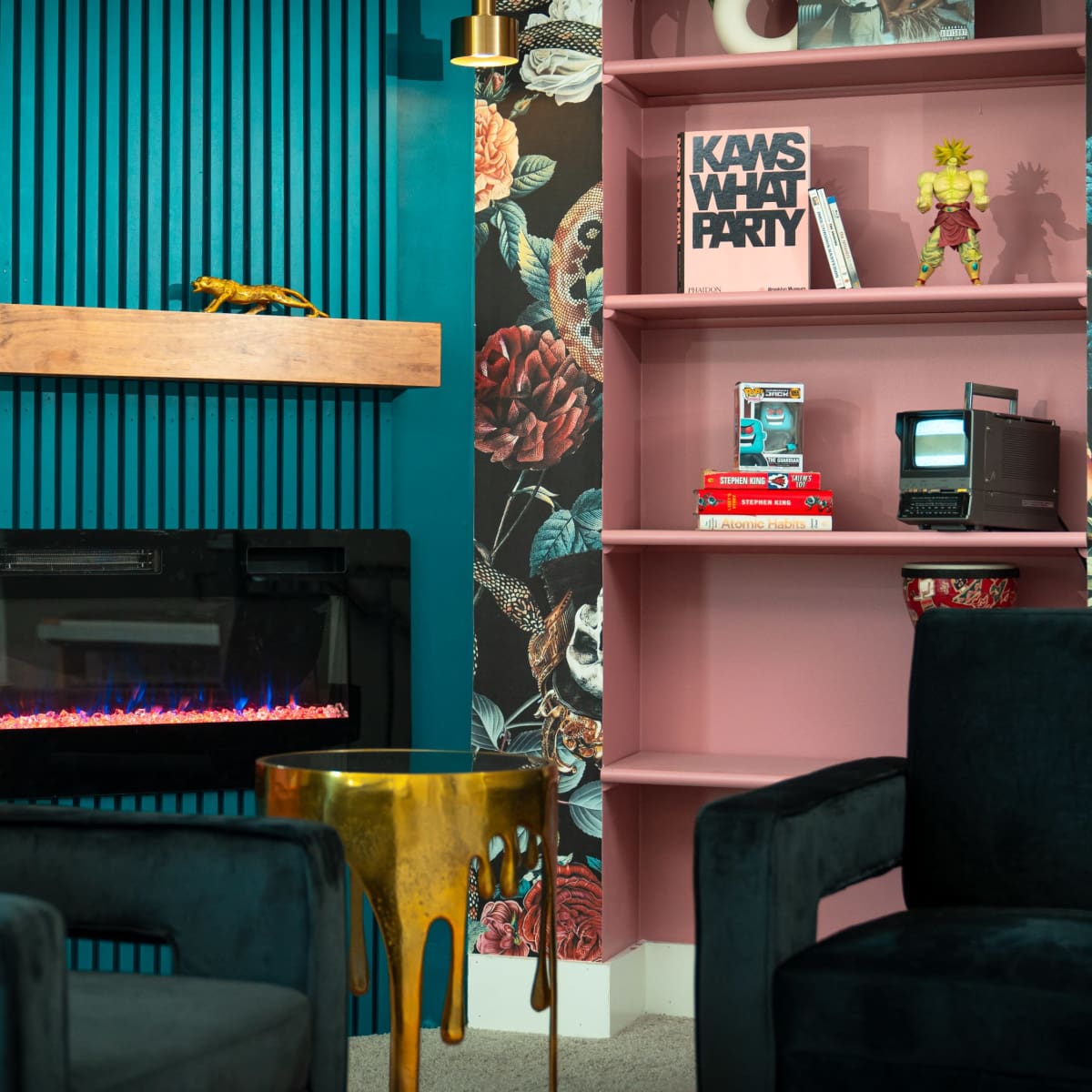 A modern living room with two velvet chairs facing a wall-mounted electric fireplace and a bookshelf featuring decorative items and a vintage television.