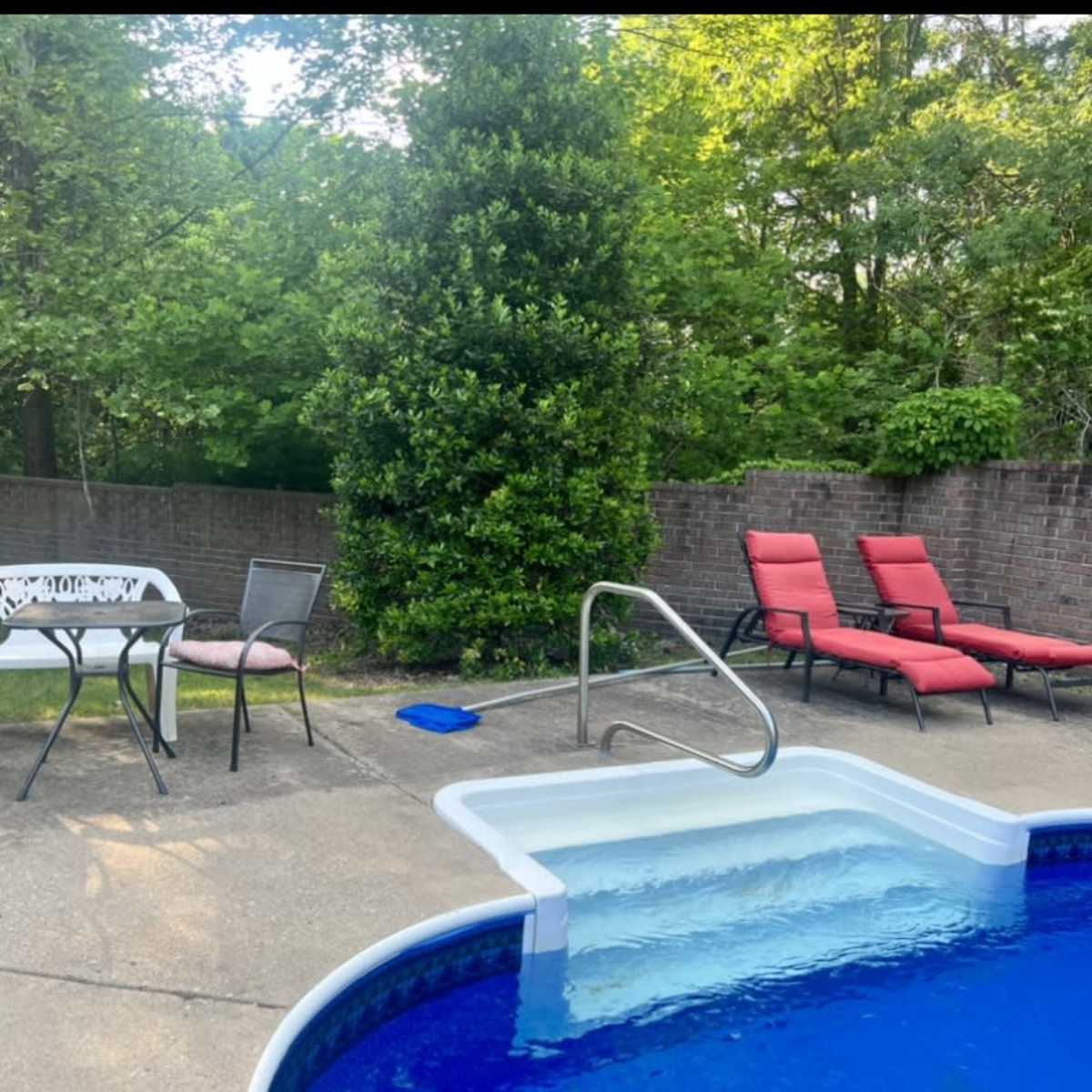 A swimming pool surrounded by two red lounge chairs, a small table with two chairs, and a landscaped area with trees in the background.