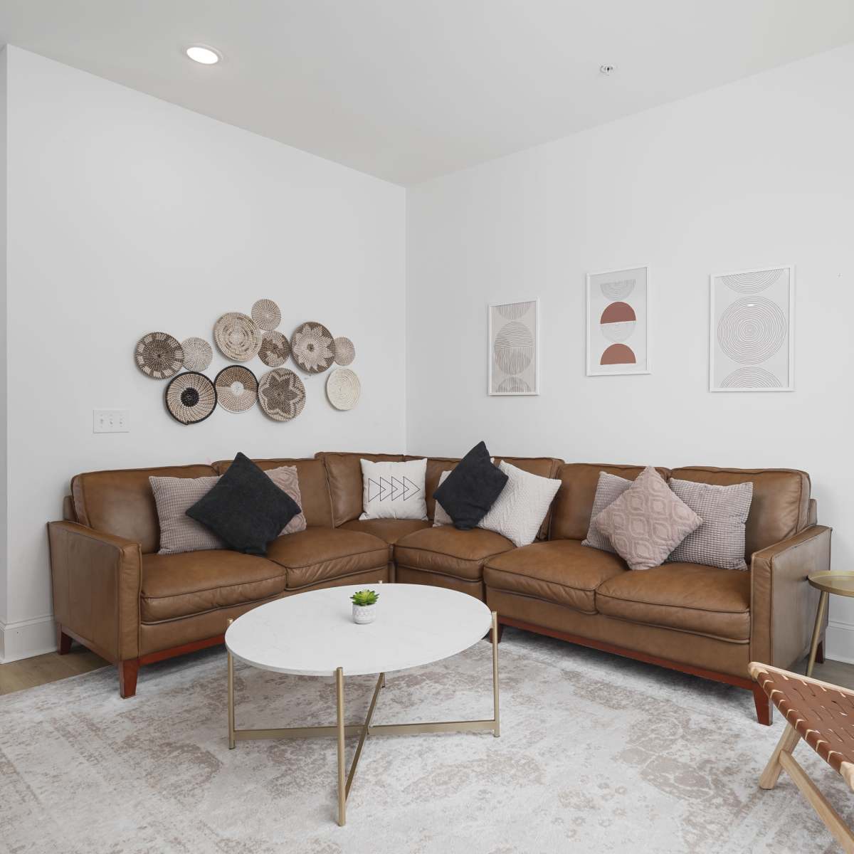 A modern living room features a brown sectional sofa with decorative cushions, a round coffee table, a floor lamp, and framed artwork on the walls.