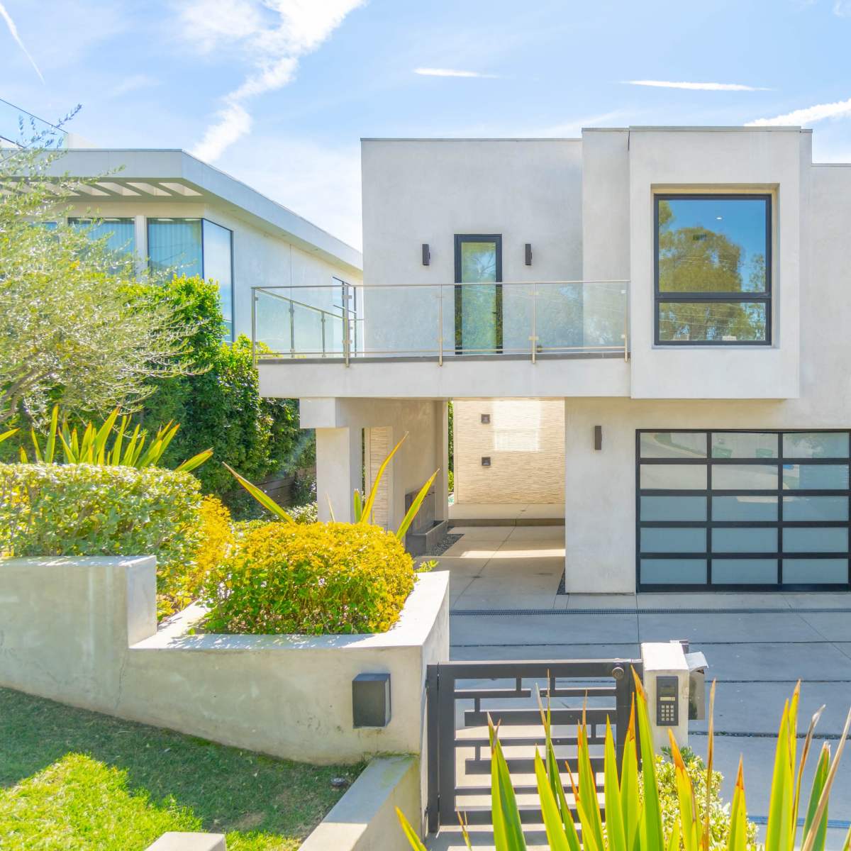 A modern two-story house with a glass-railed balcony, a large garage door, and landscaped greenery in the front yard.