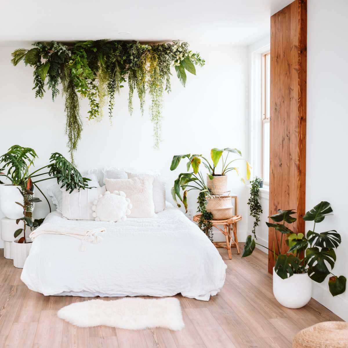 A bright bedroom featuring a white bed adorned with pillows, surrounded by various indoor plants and wooden accents.