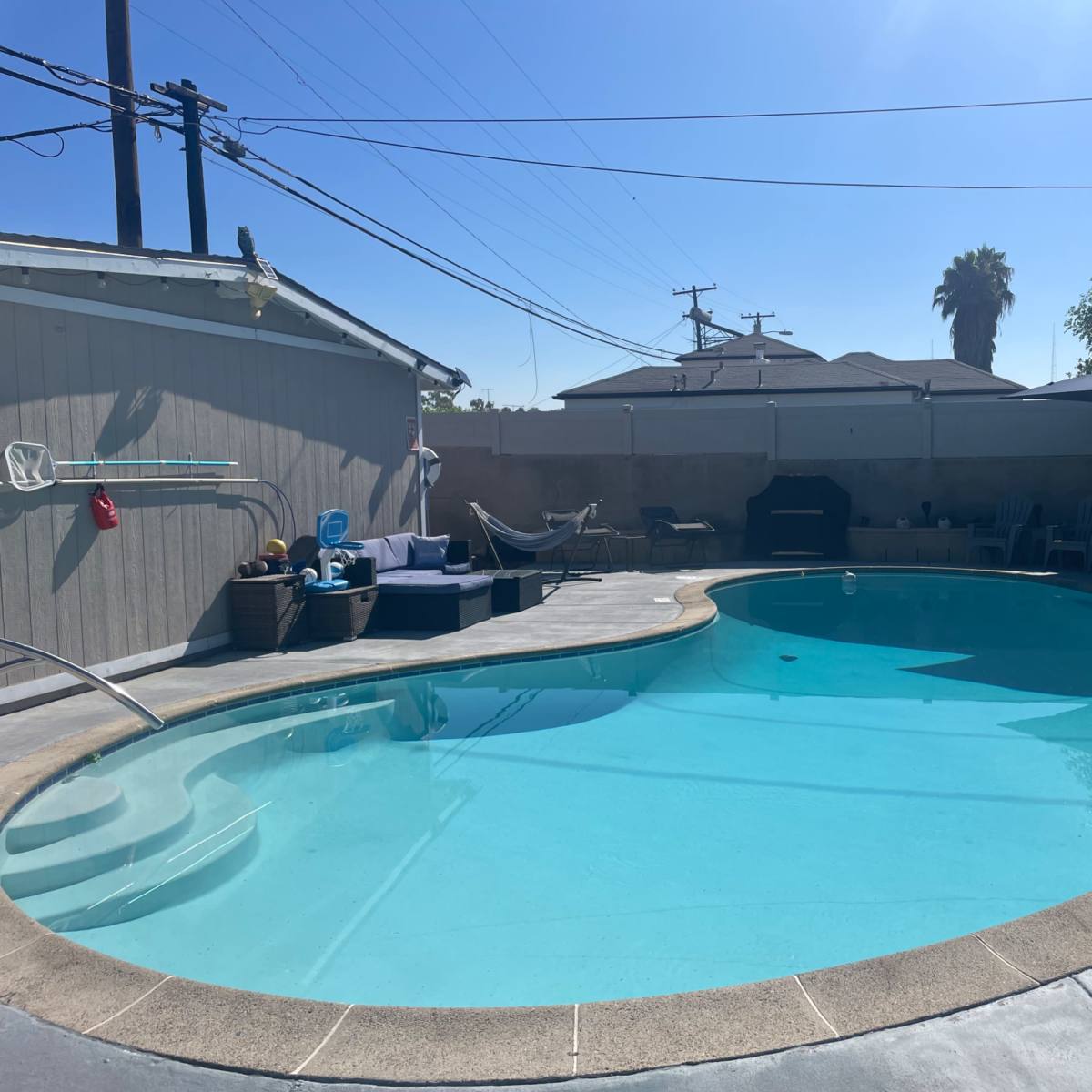 A swimming pool surrounded by a concrete patio, various outdoor seating arrangements, and residential buildings in the background under a clear blue sky.