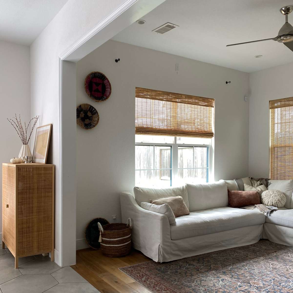 A bright living room features a white sofa with throw pillows, a wooden cabinet, and a patterned area rug on a tile floor.