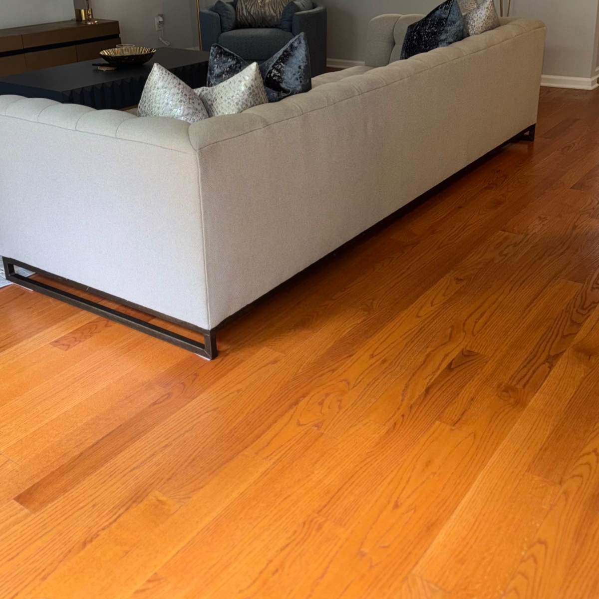 A light gray sofa with decorative pillows is positioned across a wooden floor, facing a coffee table in a modern living room that leads into a kitchen area.