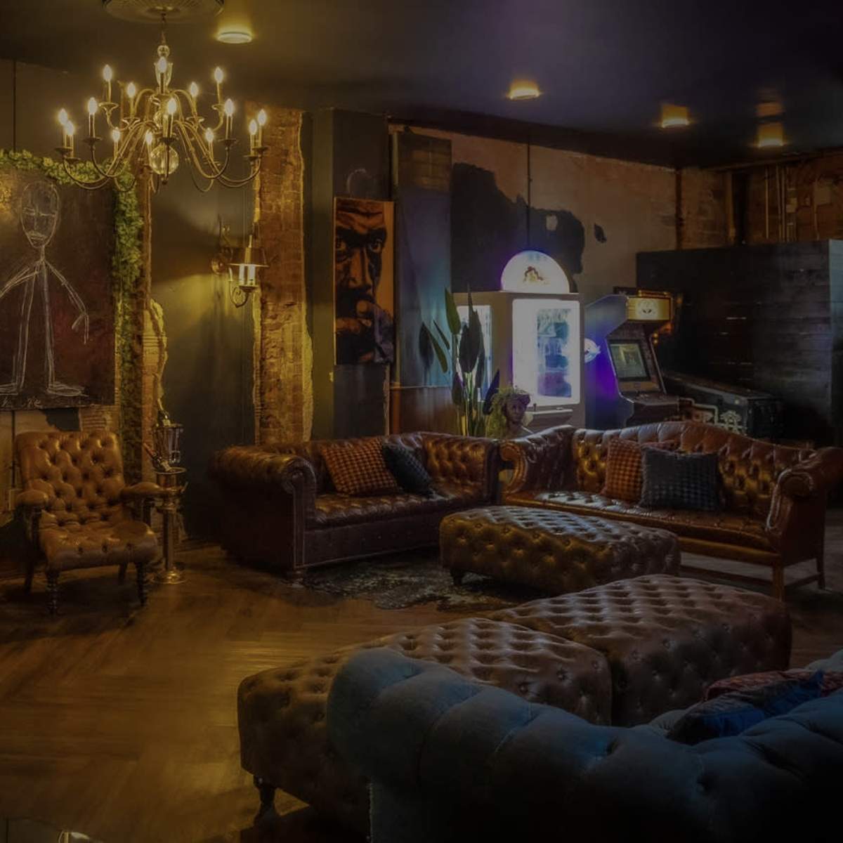 A dimly lit lounge area with leather sofas, a chandelier, and an arcade machine in a rustic setting.