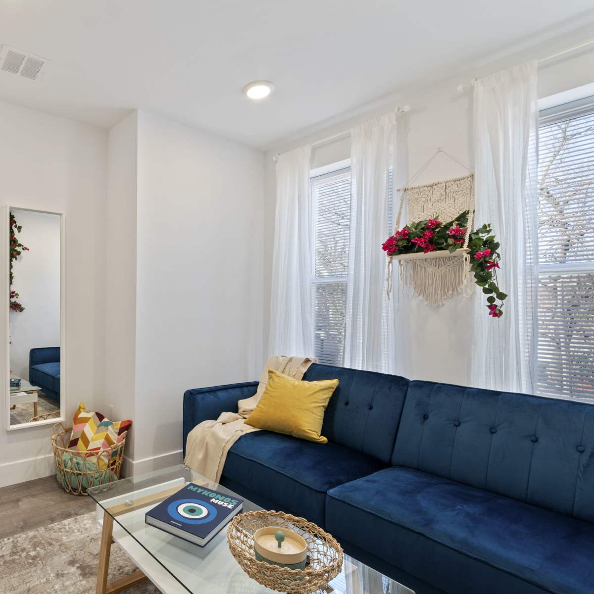 A modern living room features a blue sofa, a glass coffee table, a large mirror, and decorative plants by the windows.
