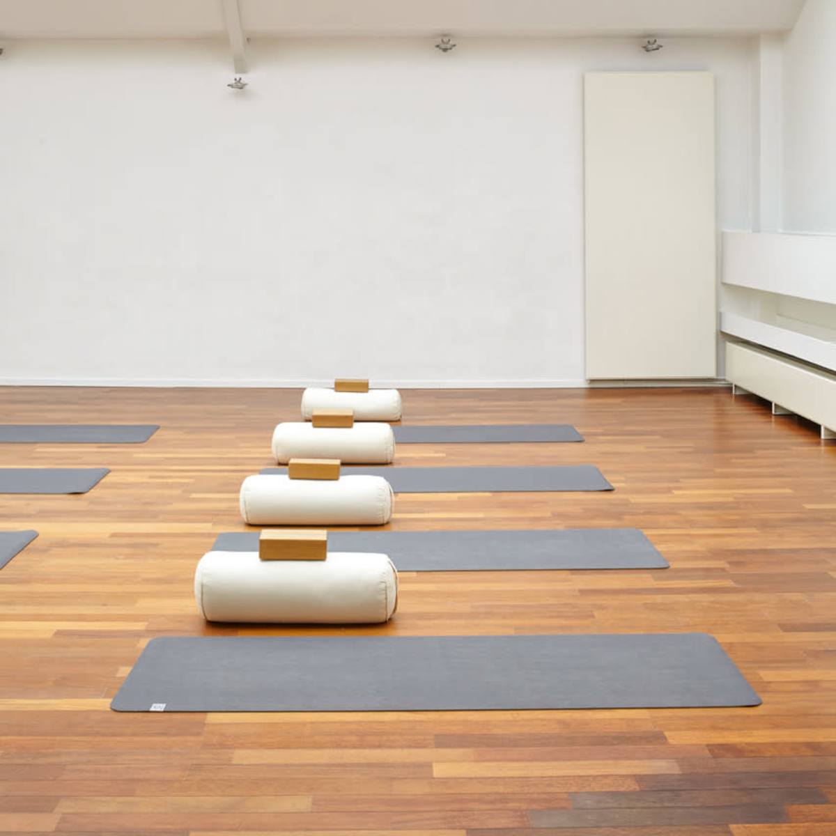 A spacious yoga studio features wooden flooring, several gray mats, and cylindrical bolsters arranged neatly in rows under a skylighted ceiling.