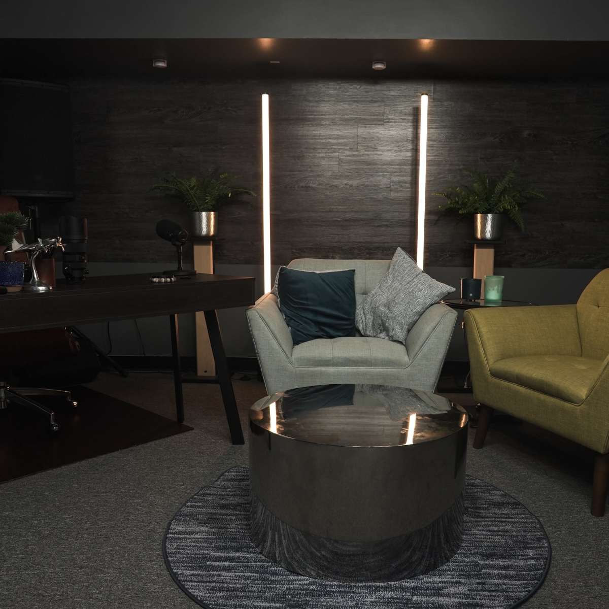 A modern lounge area with a gray armchair, a yellow chair, a round coffee table, and a desk in a dimly lit room featuring wooden accents and plants.