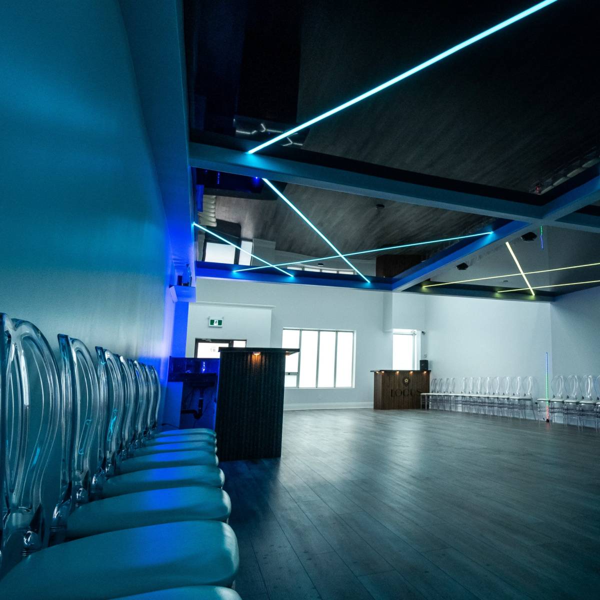 A spacious, modern room with transparent chairs arranged in rows and vivid blue and green lighting on the ceiling.