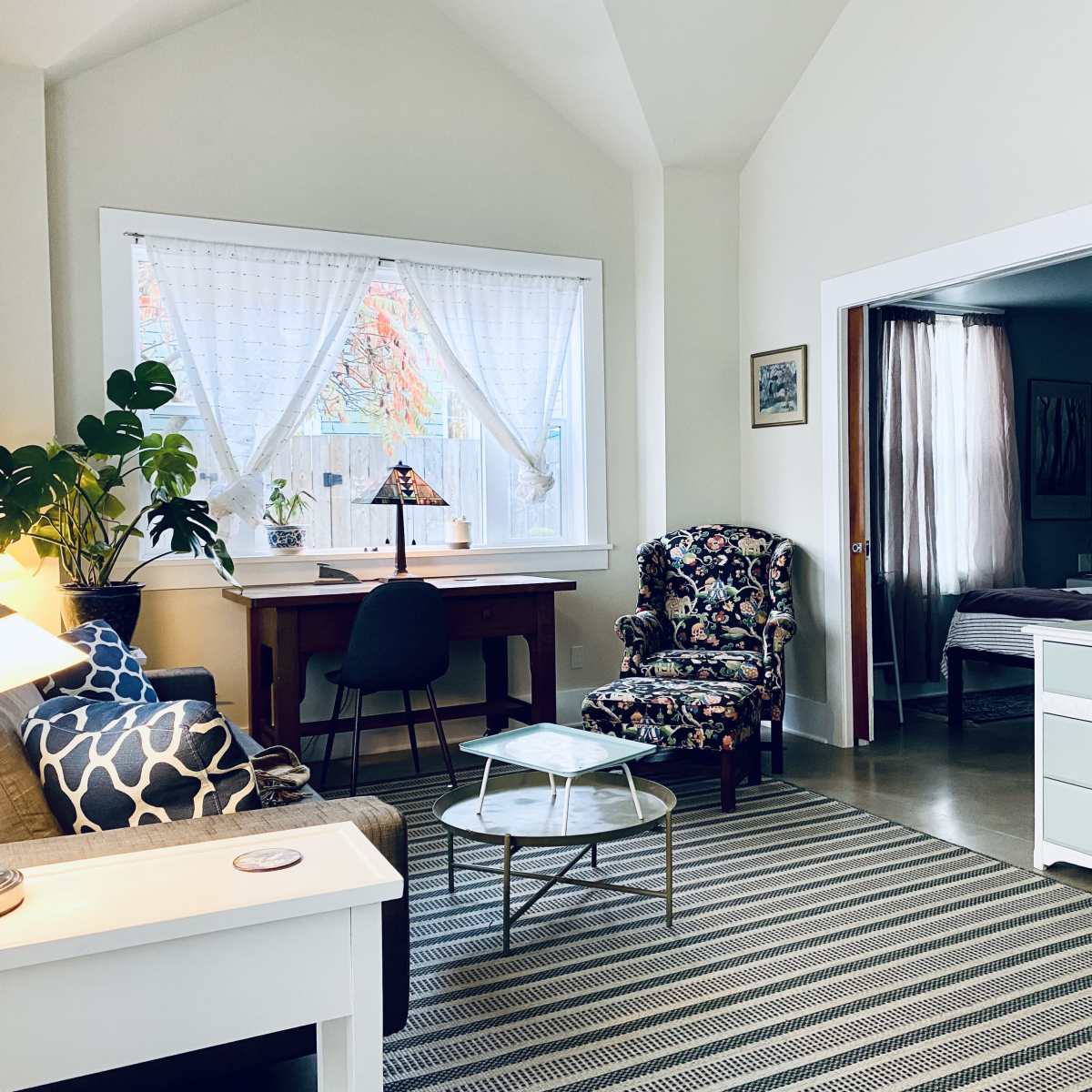 A bright living space featuring a floral armchair, a wooden desk by a window, a light-colored dresser, and a television mounted on the wall.