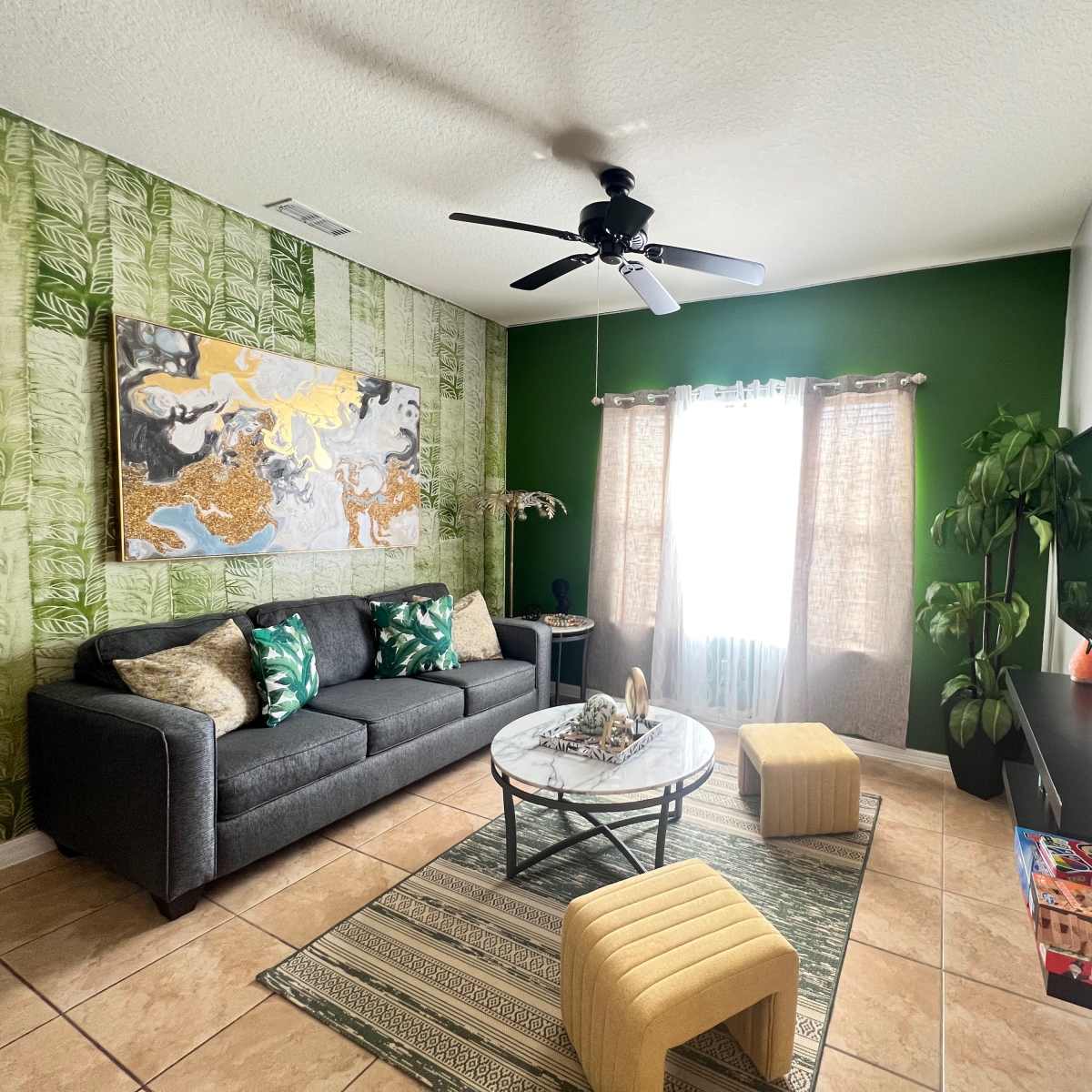 The living room features a gray sofa, a round coffee table, a large colorful wall painting, and a green accent wall with natural light coming through the sheer curtains.