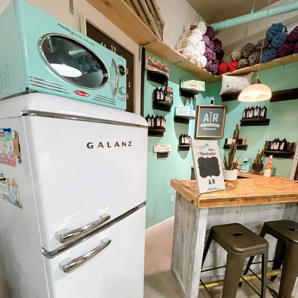 A retro-style white refrigerator next to a wooden table with metal stools in a brightly colored room adorned with shelves of yarn and craft supplies.
