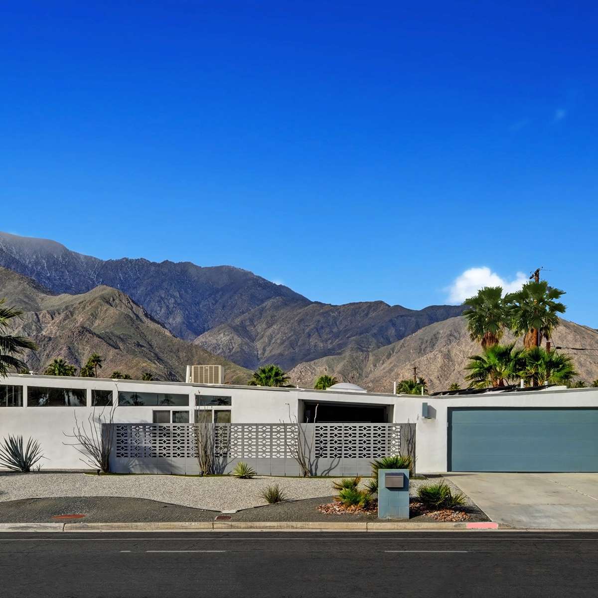 A modern white house with a gray garage is set against a backdrop of mountains and palm trees.