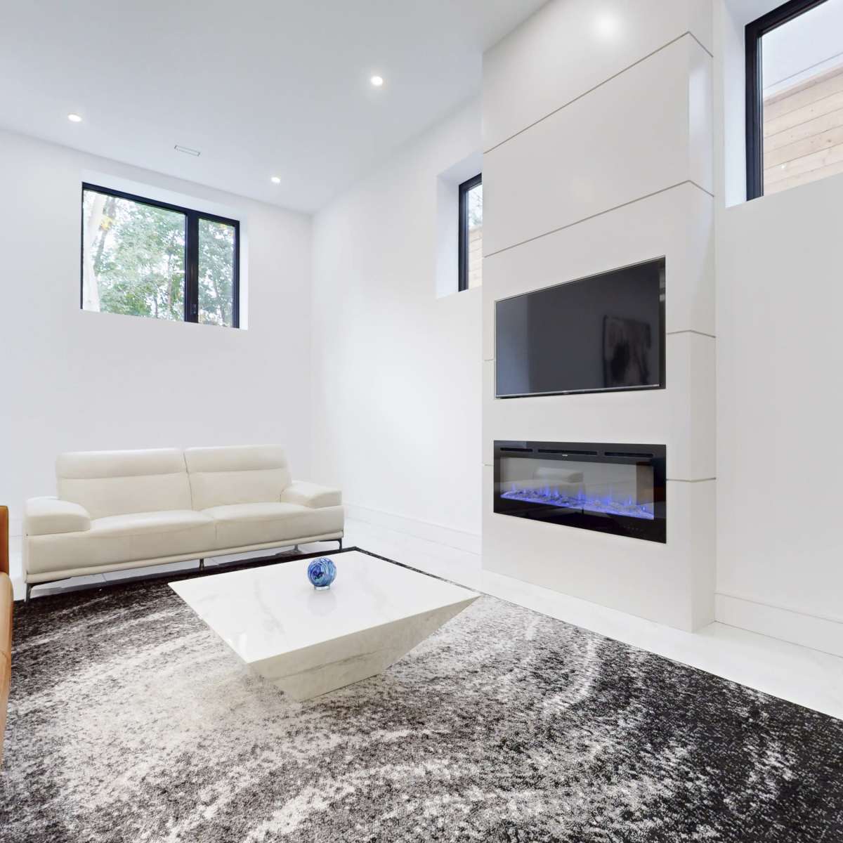A modern living room with a white wall, a beige sofa, a flat-screen TV mounted above a sleek fireplace, and a patterned black and white rug under a coffee table.