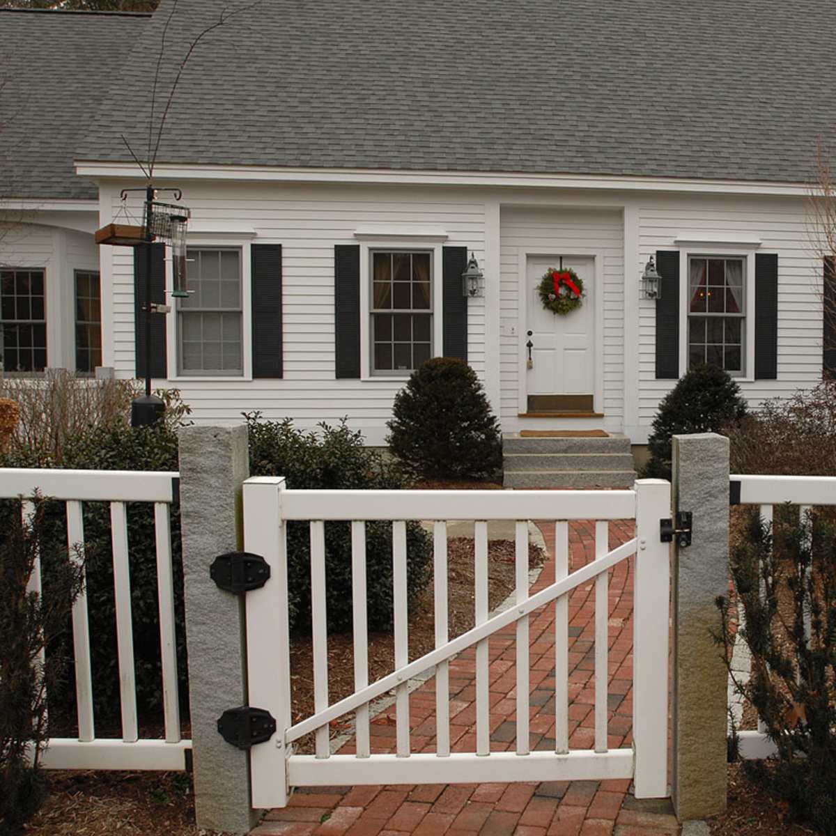 A white wooden fence with a gate leads to a house with a grey roof, featuring a wreath on the front door and landscaped gardens on either side.