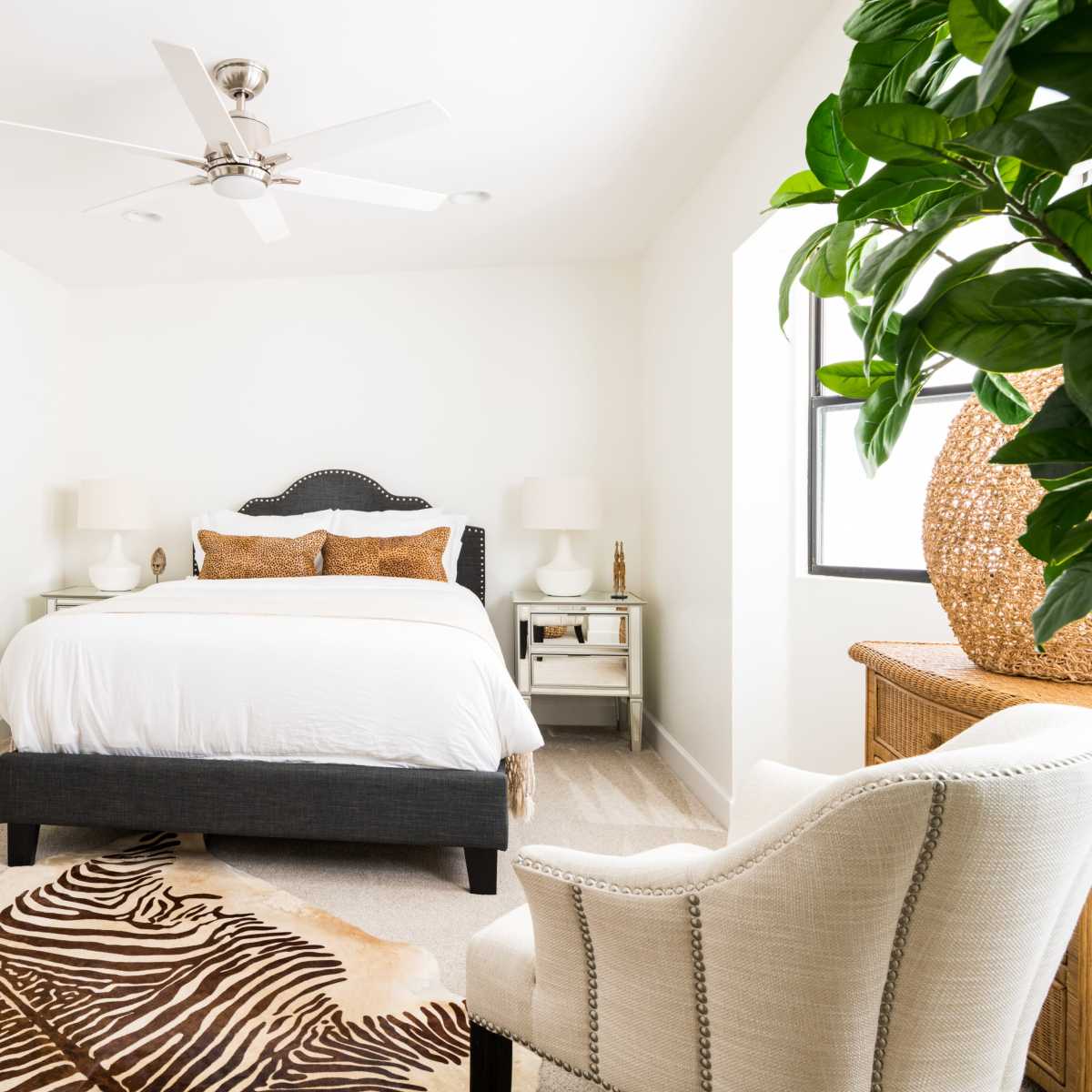 A bright, minimalist bedroom featuring a bed with white bedding, a ceiling fan, and a zebra-print rug, complemented by a potted plant and decorative furniture.