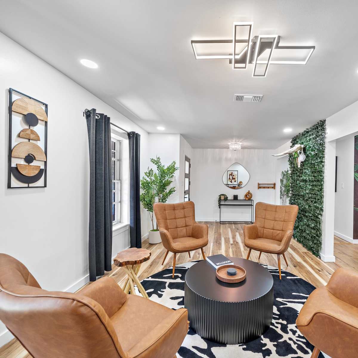 A modern living room featuring four tan chairs arranged around a black circular coffee table, with a green wall and a forest-themed mural in the background.