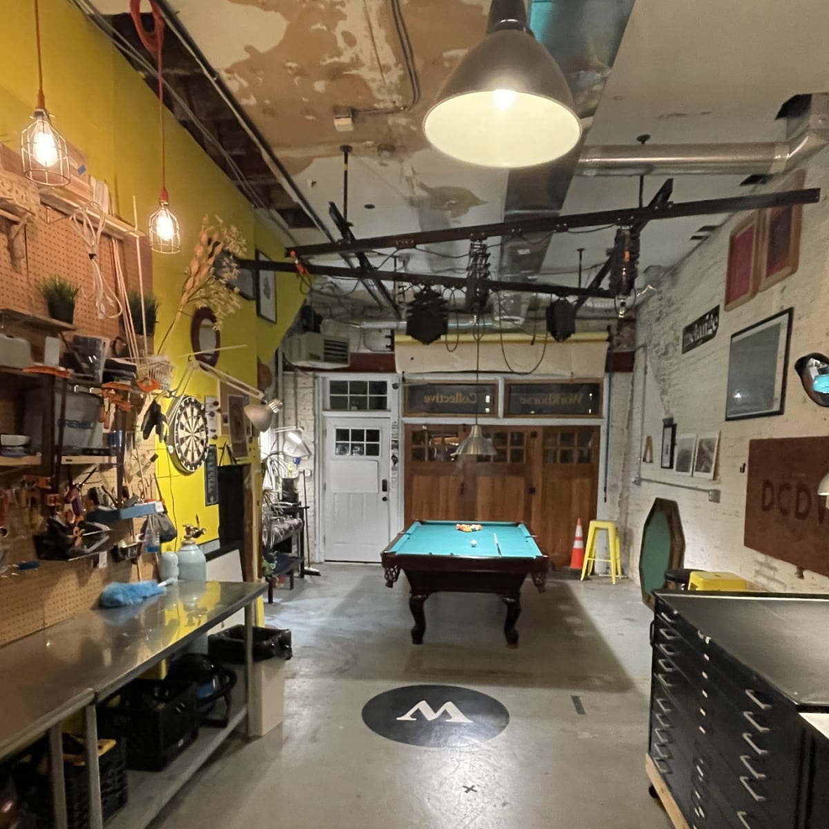 A spacious workshop or game room with a green pool table centered on a concrete floor, surrounded by various tools and equipment against the walls.