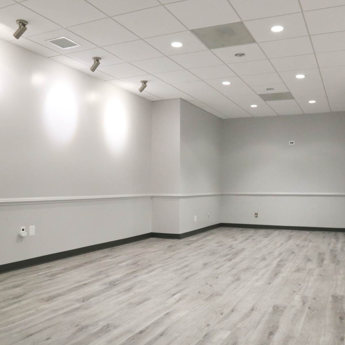 A well-lit, empty room featuring gray walls, a wood-like floor, and a simple ceiling design.
